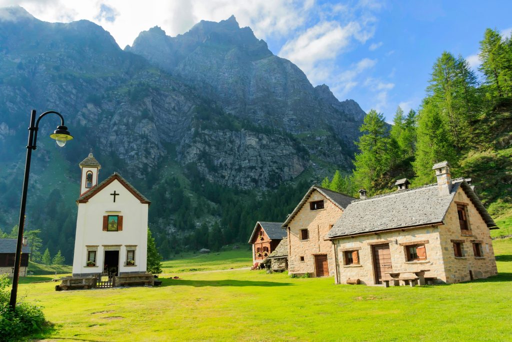 Stone cottages and a small chapel in front of Monte Rosa with lush green meadows, a scenic spot for hiking in Italy.
