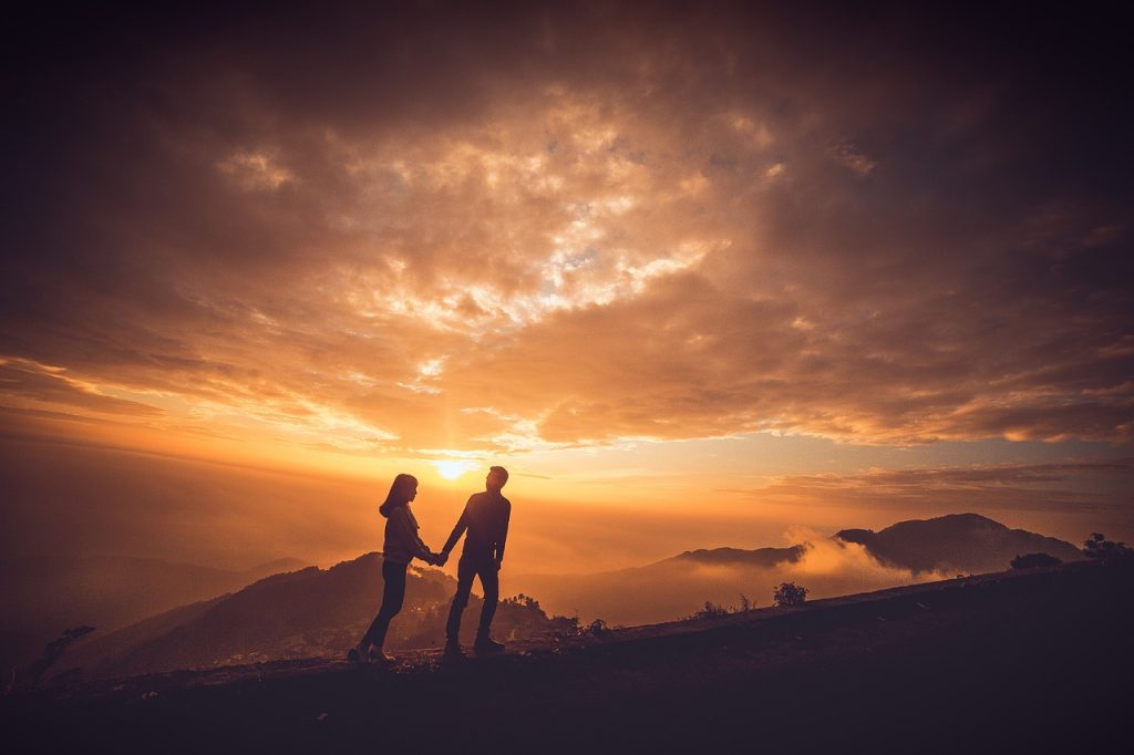 Couple holding hands while hiking on a mountain during a sunset, surrounded by scenic views and cloudy skies.