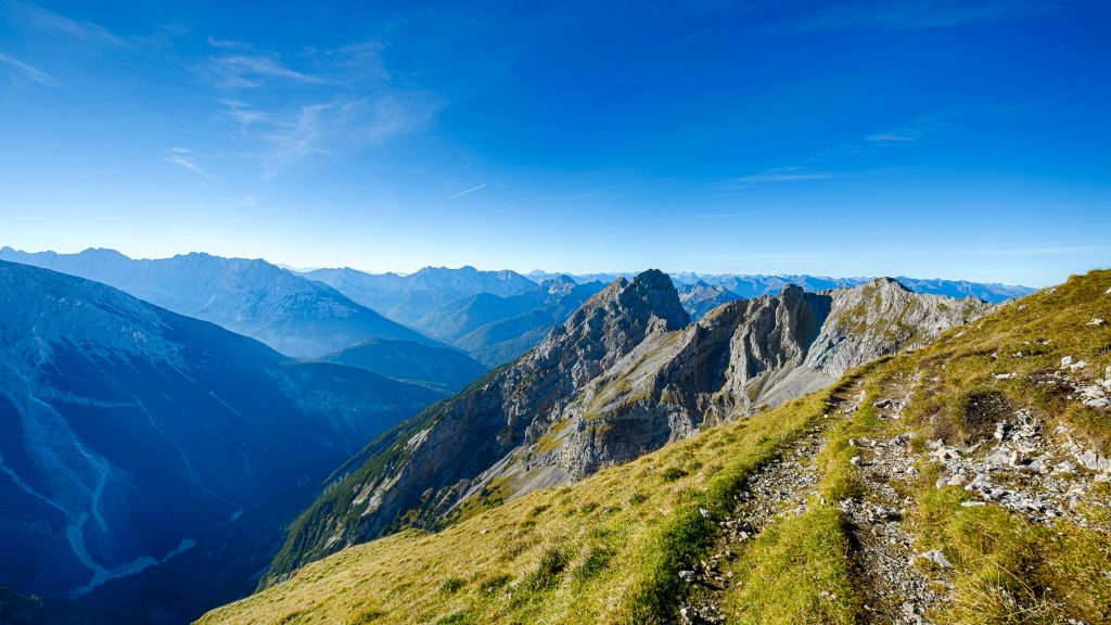 Scenic view of the German Alps with rugged peaks, green slopes, and a winding trail under a clear blue sky.