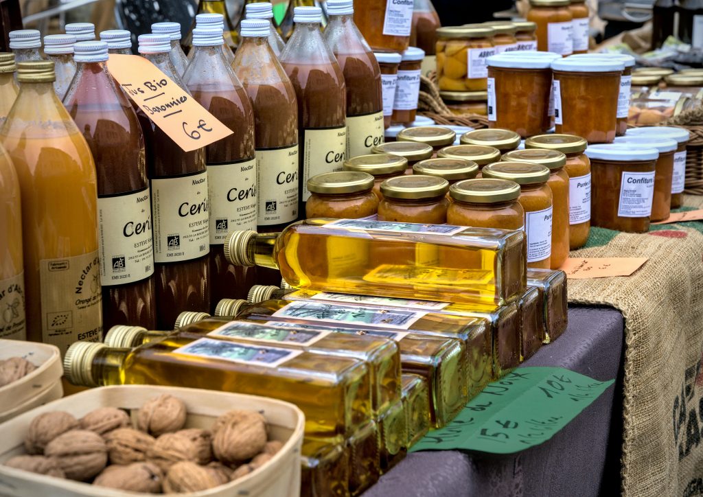 Bottles of honey, jam jars, and natural oils displayed at a market alongside walnuts and price tags for homegrown foods.