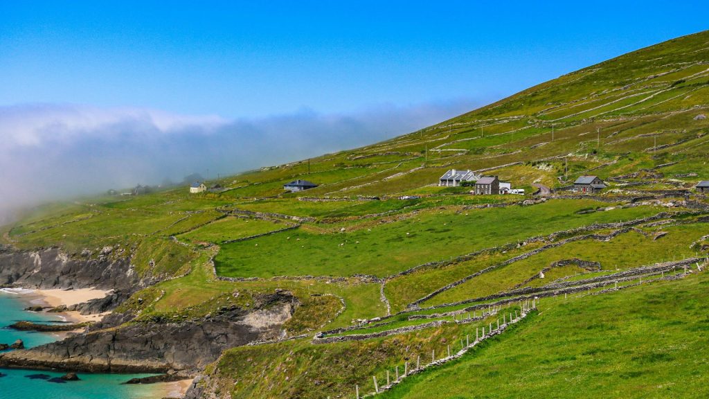 A scenic hillside along The Dingle Way with stone walls, small houses, and a rugged coastline with turquoise waters.