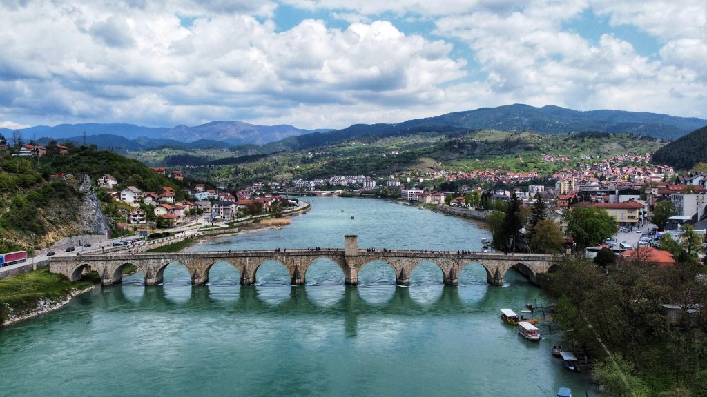 Historic stone bridge over a river in Bosnia, surrounded by lush hills and town views, perfect for outdoor adventures.