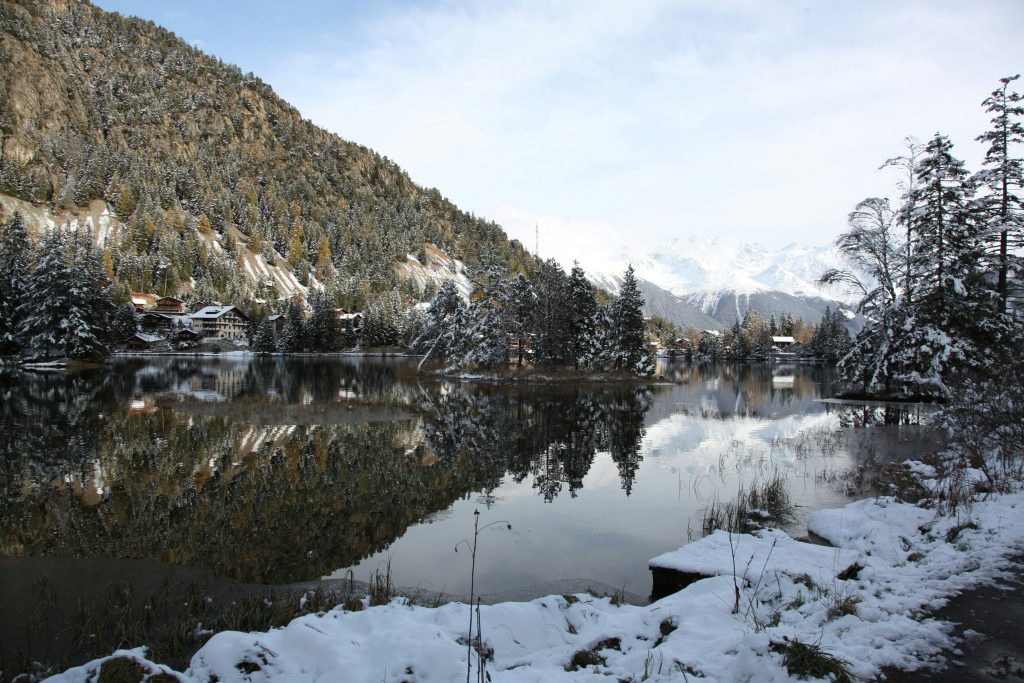 Snowy Champex-Lac with mountain reflections in the lake, surrounded by pine trees, along the Mont Blanc hiking route on a crisp winter day.