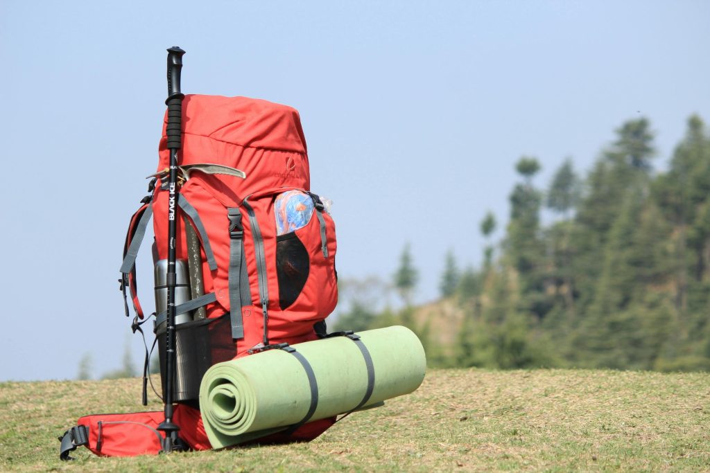 Red hiking backpack with trekking pole, green rolled mat attached, and a natural outdoor backdrop, ideal for hiking and camping.