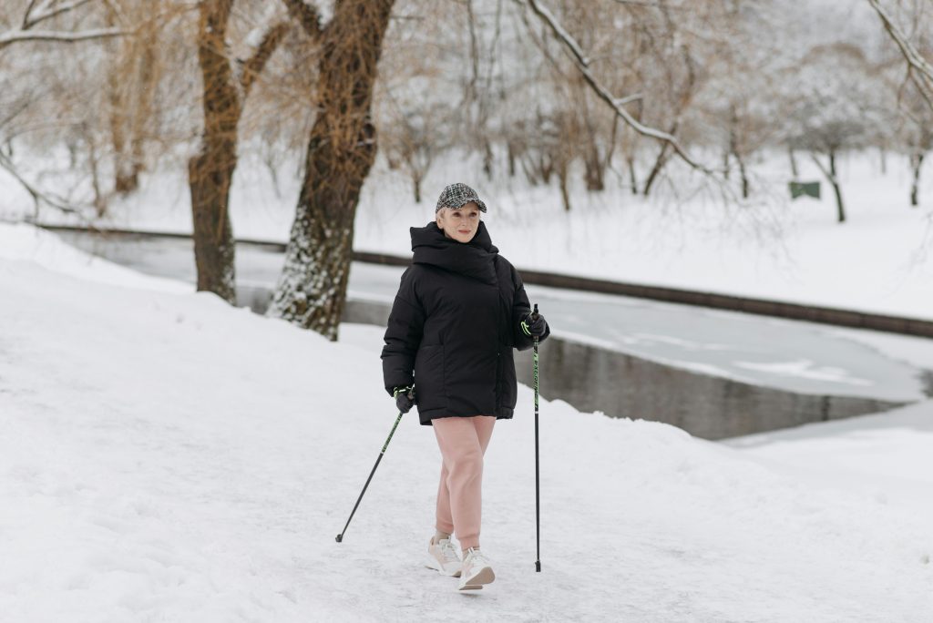 A woman walking through the snow with trekking poles, wearing a warm jacket and pants, with a snowy landscape and frozen river in the background.