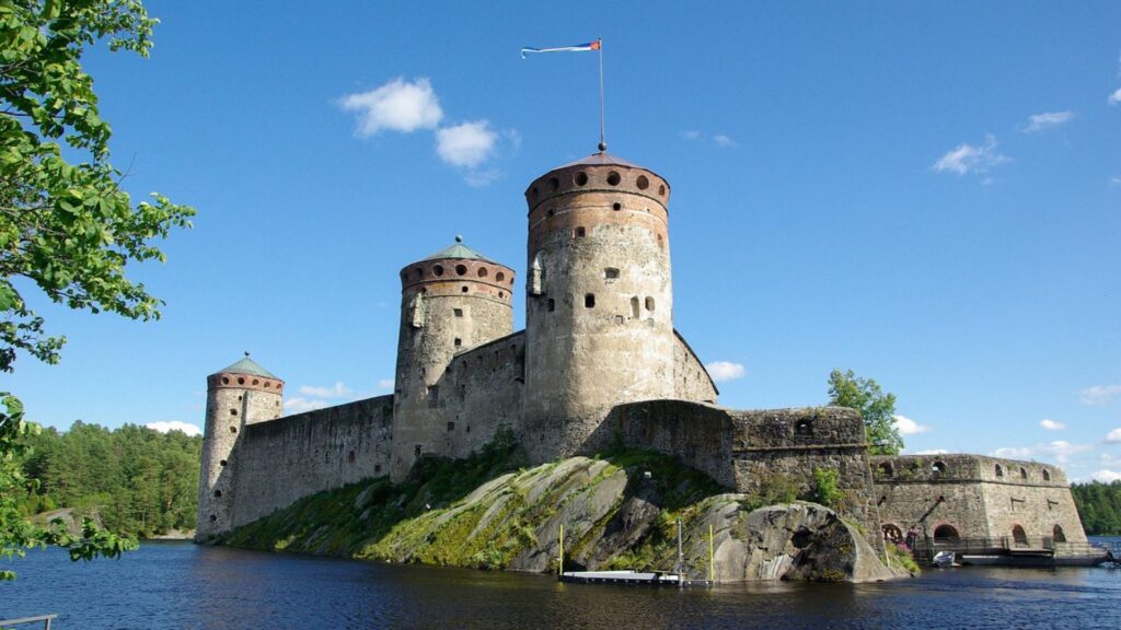 Historic Olavinlinna Castle surrounded by water and lush greenery under a clear blue sky, a scenic landmark often visited during hiking in Finland adventures.