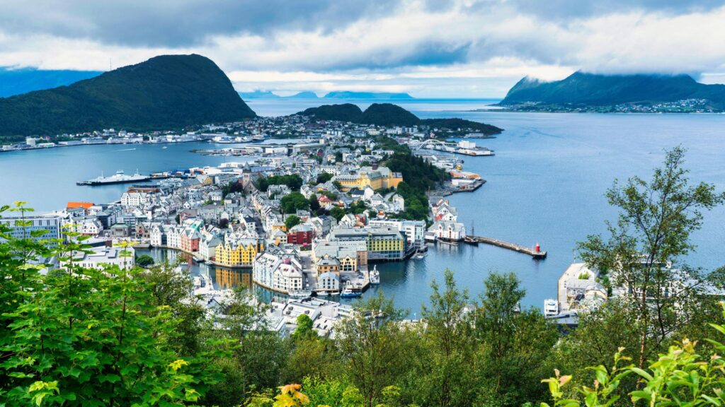 Panoramic view of Ålesund, Norway with colorful buildings, harbor, and surrounding mountains under cloudy skies, seen from a lush hillside viewpoint.