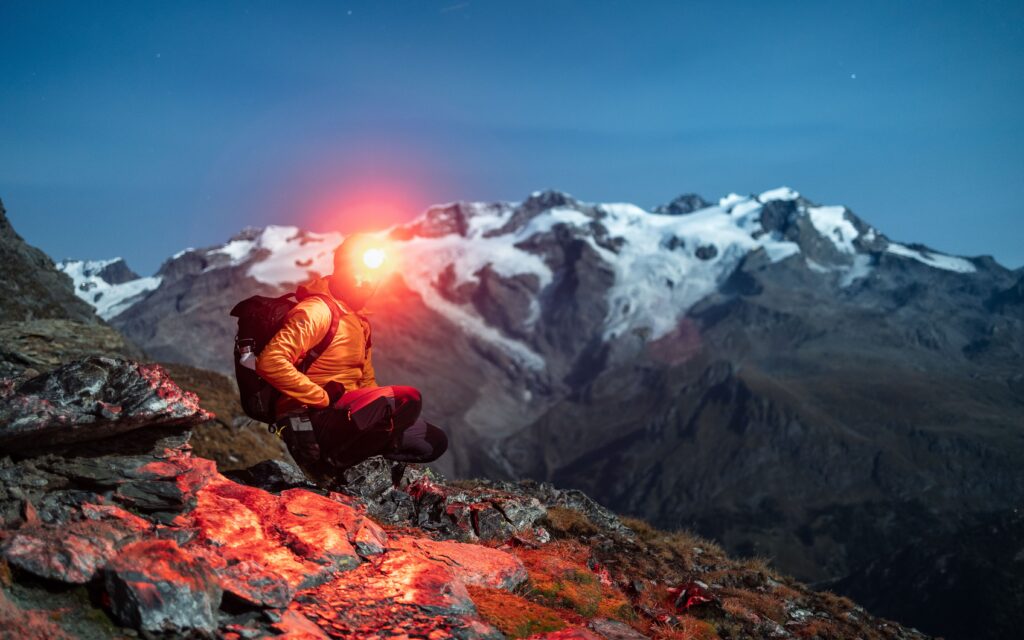 How to Choose a Headtorch: hiker wearing a bright headtorch on a rocky alpine trail before sunrise with snowy peaks behind.