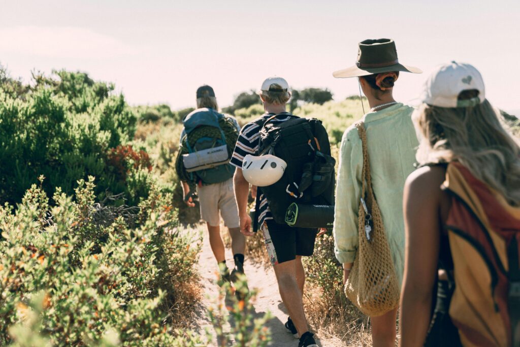Group of hikers on a sunny trail with backpacks and hats, enjoying warm weather and summer hiking adventures.