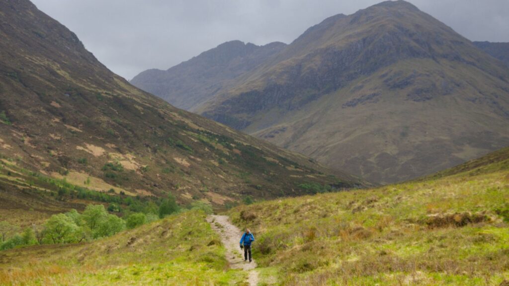 Hiker walking through dramatic mountain scenery on the Cape Wrath Trail in the Scottish Highlands under cloudy skies.