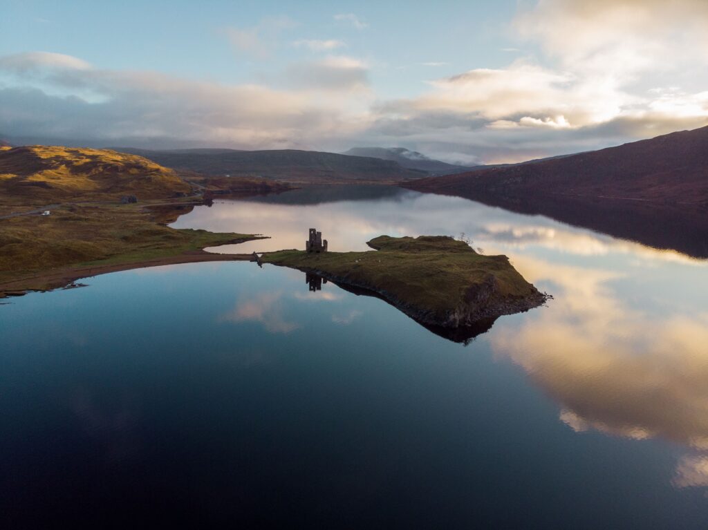 Aerial view near Inchnadamph featuring a tranquil loch, historic castle ruins, and rugged Highland scenery in Scotland.