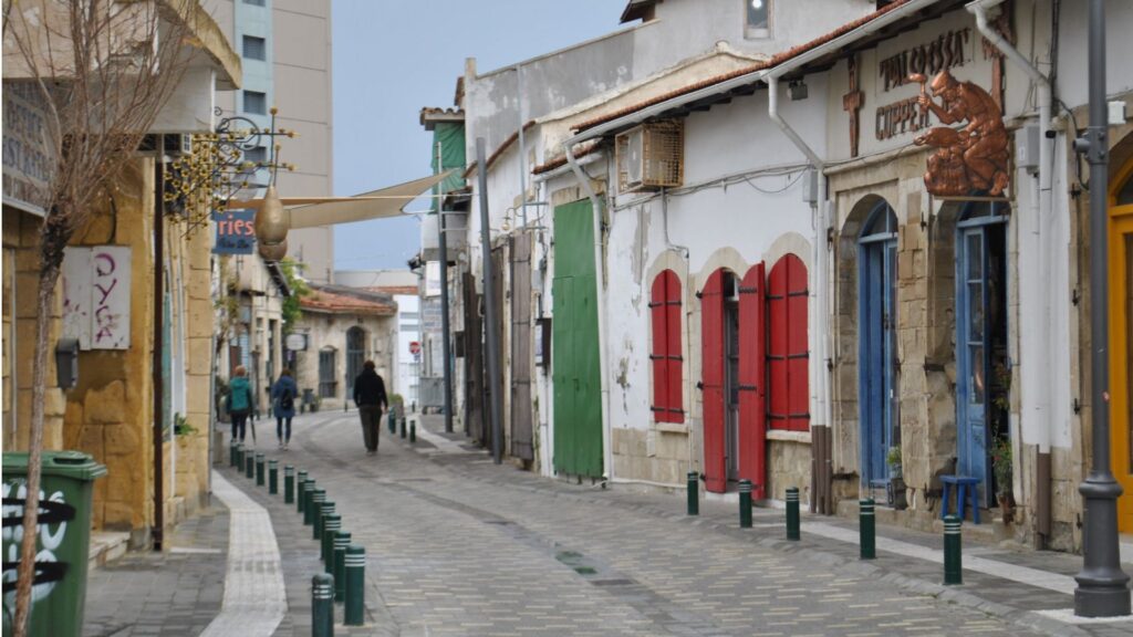 Narrow pedestrian street in a Mediterranean town with colorful shutters and older stone buildings; a few people walk in the distance.