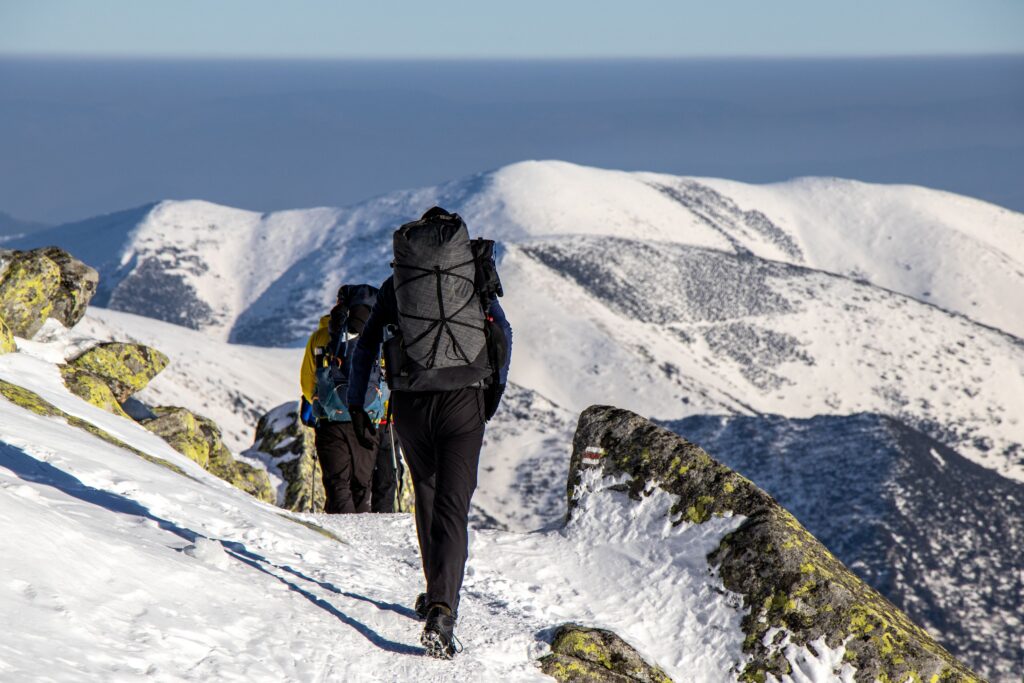 Hikers trekking snowy mountain trail on long day hikes with backpacks and alpine views in winter wilderness.