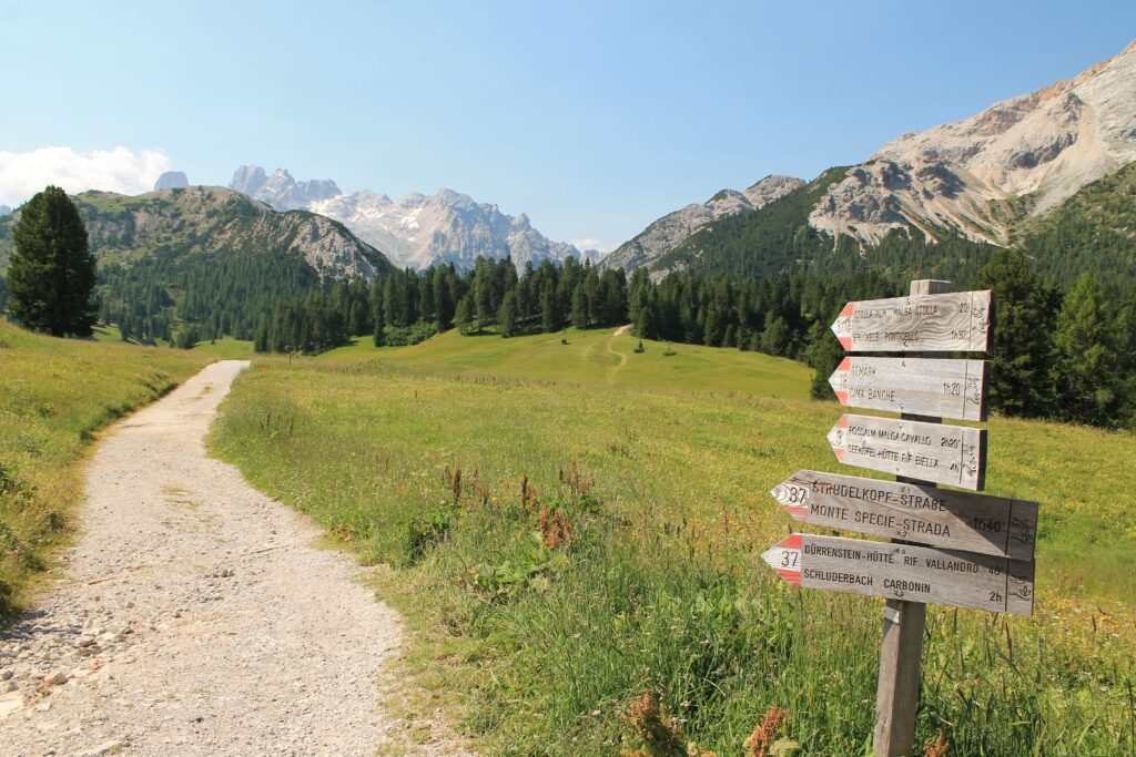 routeplainandsimple Kayde Parker Gravel hiking path through a green meadow toward distant mountains with a wooden signpost on the right showing trail directions under a clear blue sky