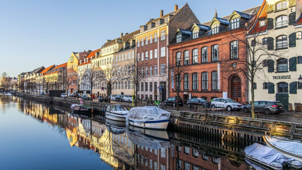 Row of colorful brick townhouses along a canal with boats moored and their reflections shimmering in the water