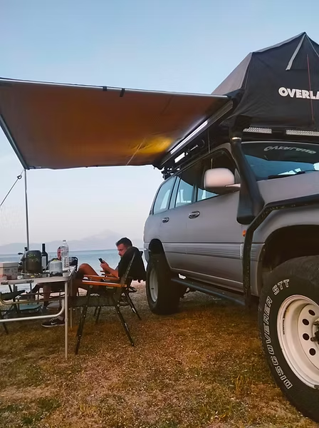 SUV with a roof top tent and a large awning set up for camping table and chairs nearby with a person sitting and using a phone beside a lake
