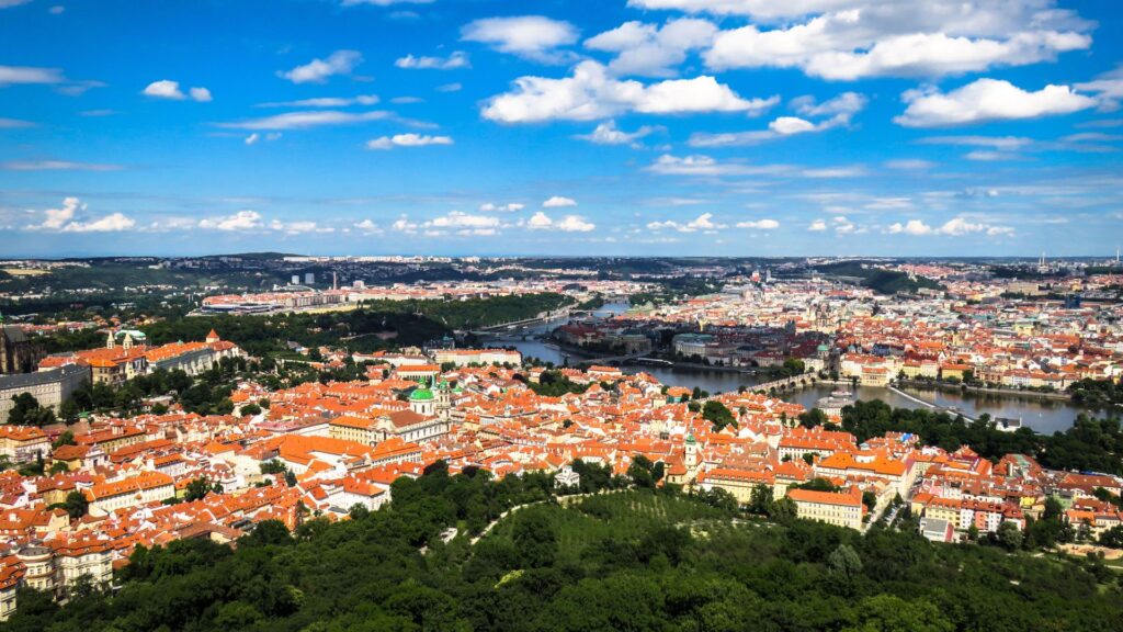 Aerial view of a historic European city with red tiled rooftops a winding river and green hills under a blue sky with clouds