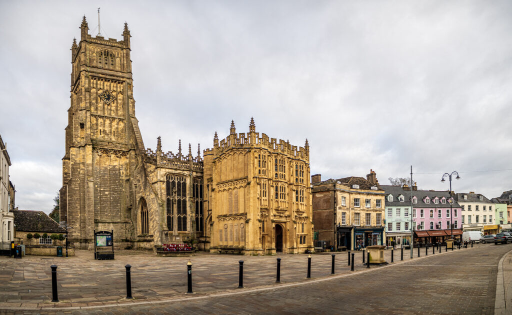 Stone Gothic church with a tall square clock tower and ornate buttresses beside a row of pastel colored townhouses in a wide town square