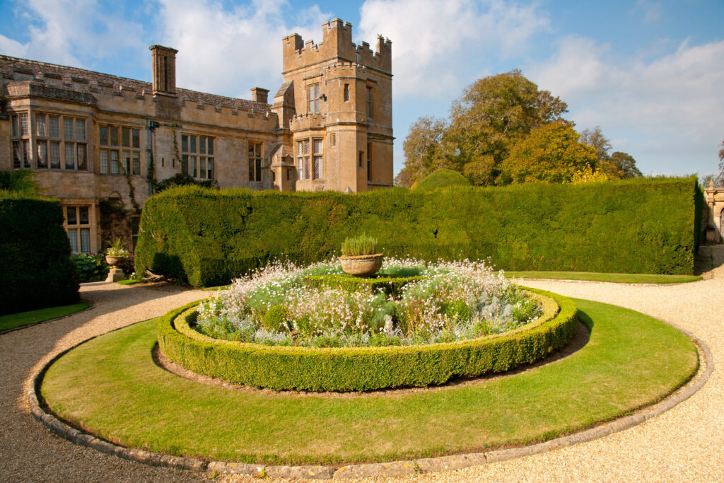Historic estate with a crenellated tower and stone wings set behind a formal circular hedged garden and gravel paths