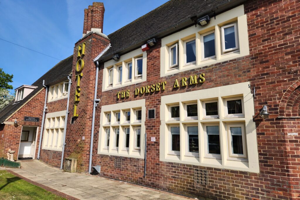 Wallsend Accom Kayde Parker Red brick pub facade with yellow lettering reading'THE DORSET ARMS' and a blue sky above, windows along cream trim, and a sign saying 'Hotel Entrance' near the door.