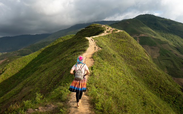 Hiker wearing colorful clothing walking along a narrow dirt trail on a green mountain ridge under a cloudy sky (back view).