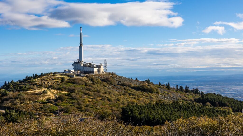 Telecommunications tower and support building perched on a grassy hillside with a blue sky and distant valley in the background