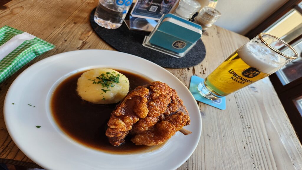 Plate with mashed potatoes in gravy and a crispy fried chicken piece on a white plate at a rustic wooden table a glass of Löwenbräu Keller beer nearby