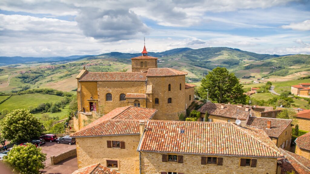 Stone buildings with red tiled roofs sit on a hillside village green fields rolling into the distance under a cloudy sky