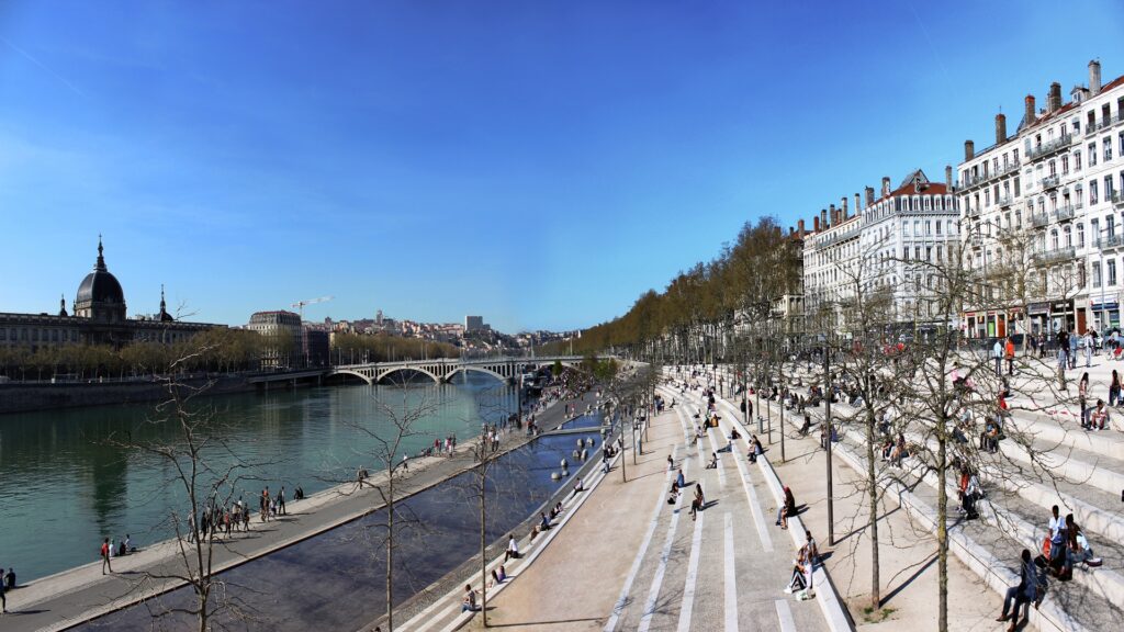City riverside with a wide pedestrian promenade people sunning and walking along the bank historic buildings line the right a bridge spans the river in the distance under a clear blue sky
