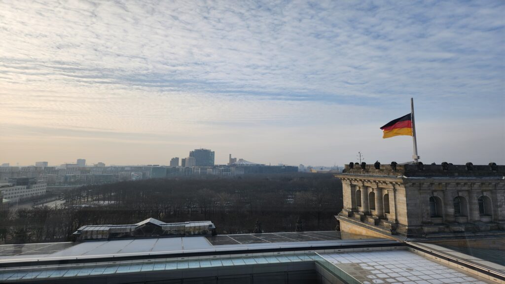 Historic building with a German flag on a flagpole in the foreground overlooking a broad city skyline under a partly cloudy sky